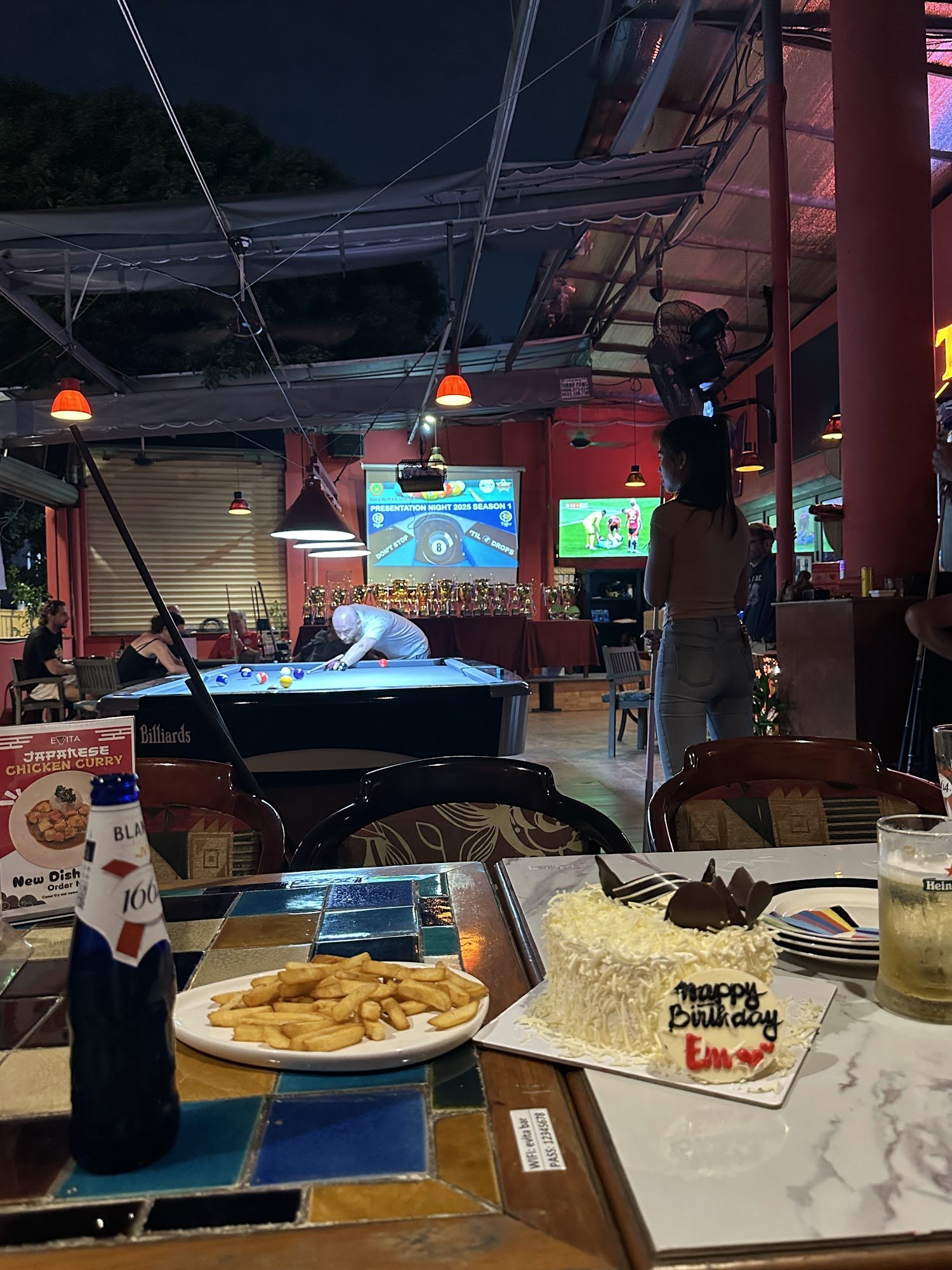 The image shows an indoor setting of a casual dining and entertainment venue during nighttime. There is a pool table in the center, with several peopl
