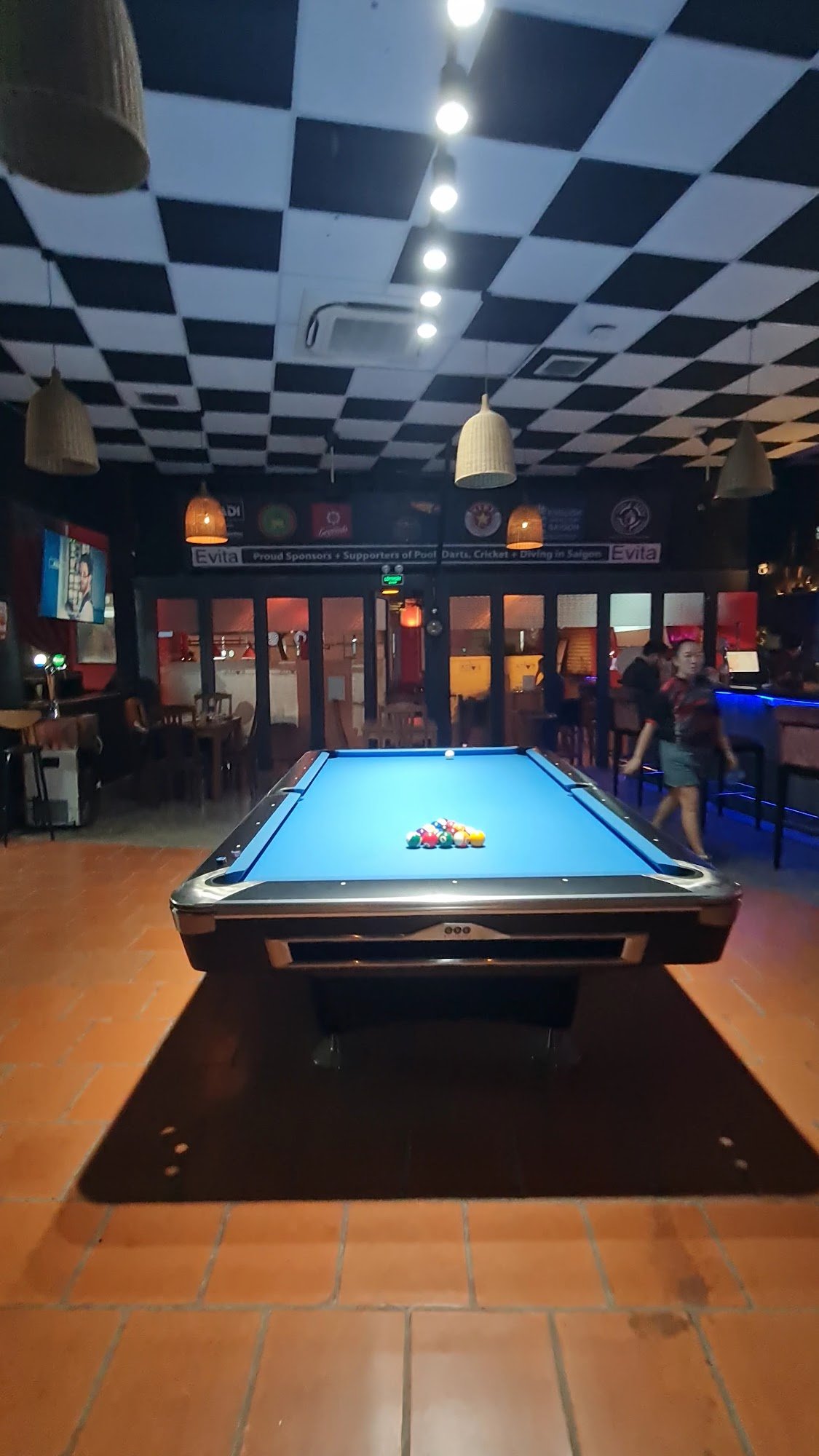The image shows an indoor pool table in a bar or pub setting. The ceiling is checkered with black and white squares, and the room has dim lighting enh