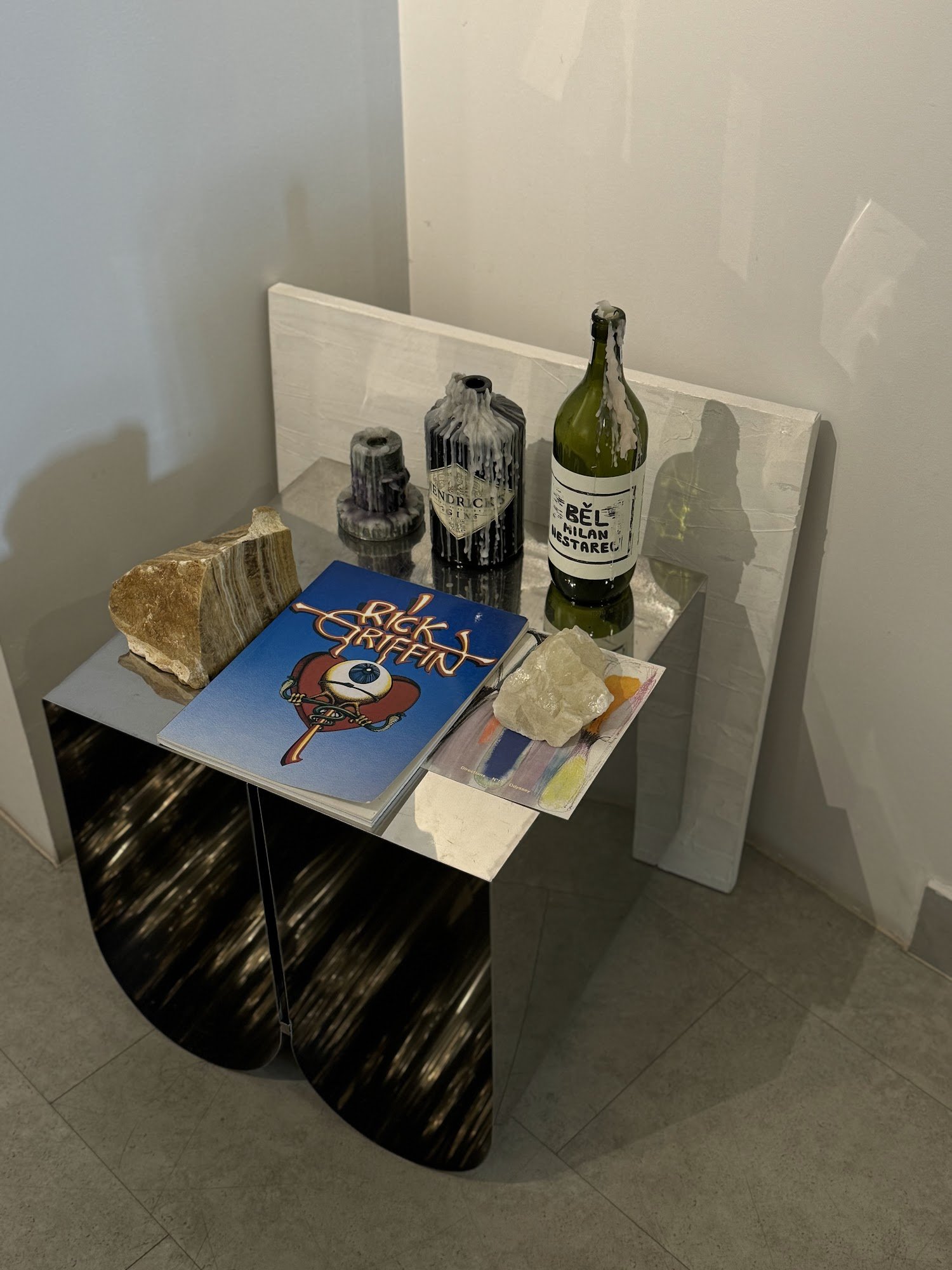 The image shows a modern interior setting featuring an artistic arrangement of various objects on a reflective surface against a plain wall.