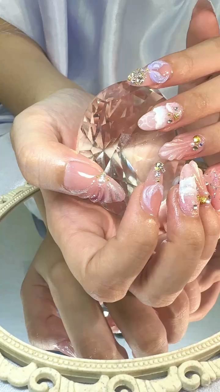 A close-up photograph of a person's hands displaying intricately designed nail art and holding a large pink gemstone in front of a mirror with an orna