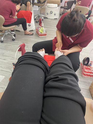 The image shows a person receiving a pedicure in what appears to be an indoor salon setting with multiple chairs and foot spa equipment visible.