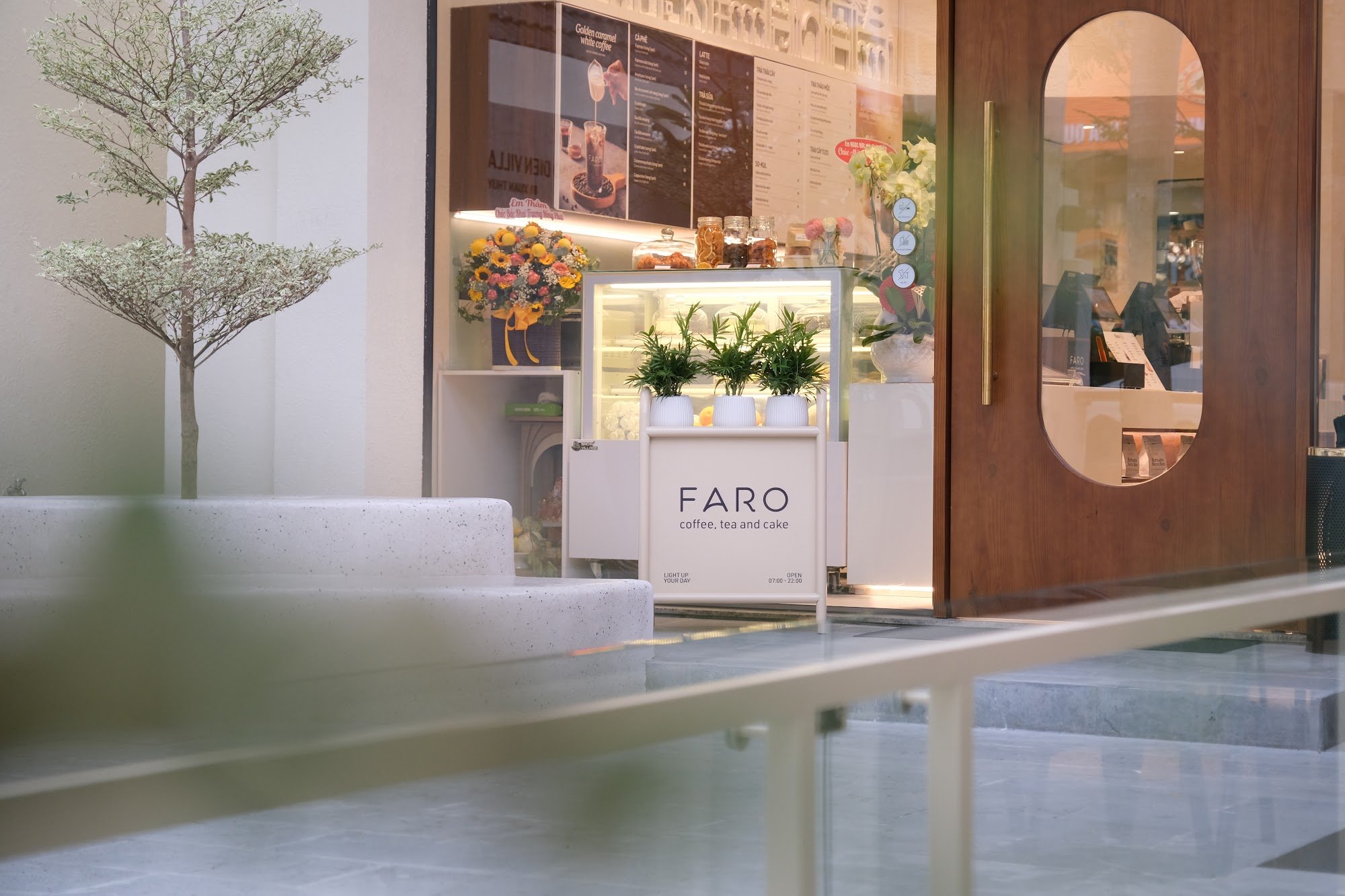 The image depicts the exterior of FARO coffee shop in Thao Dien, Saigon. It features a modern storefront with a wooden door featuring an oval window, 