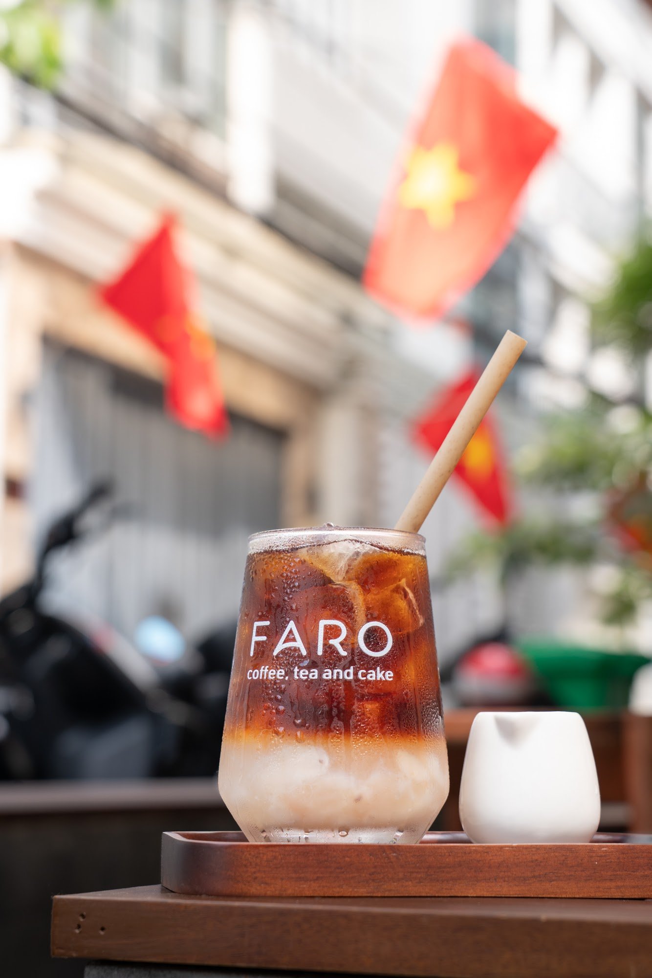 A glass of coffee from a cafe named FARO is placed on a wooden tray in the foreground with a blurred background featuring red flags, possibly indicati