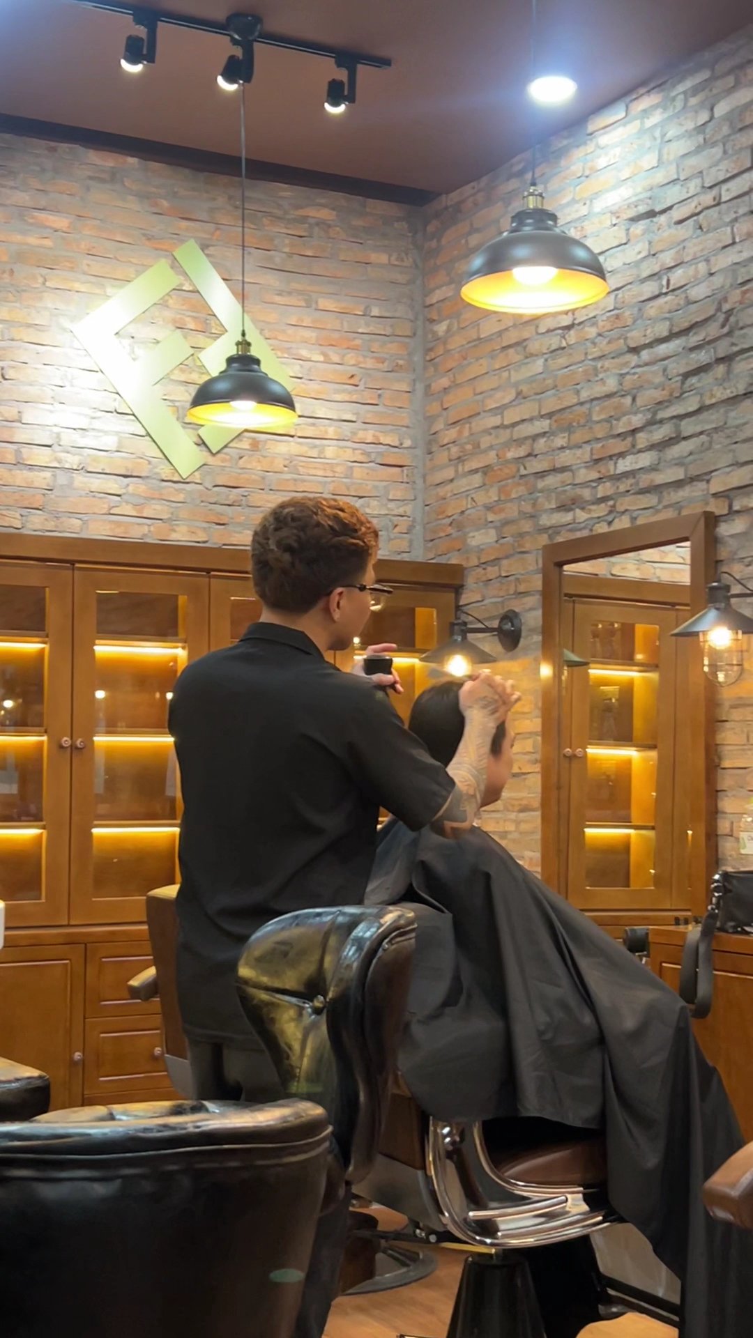 The image depicts an interior view of a barber shop or hair salon. The setting features exposed brick walls with wooden accents, including shelving an