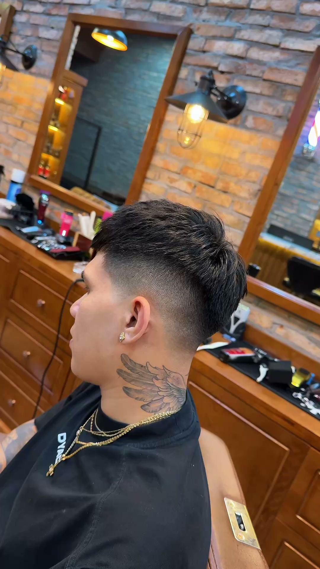 The image shows a person getting their haircut in what appears to be an interior setting of a barbershop or hair salon, with brick walls and various g