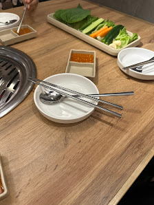 A wooden table in a restaurant setting with a built-in grill on the left, a white bowl containing chopsticks and spoons, small square dishes with sauc
