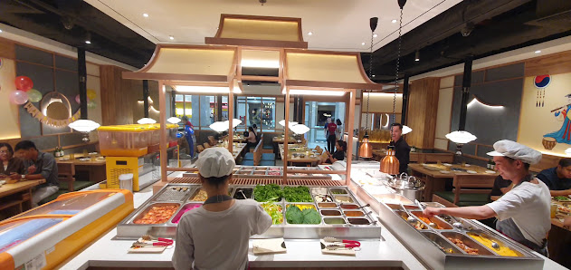 The image depicts an indoor dining area of a restaurant or food court featuring a buffet-style serving counter with various dishes displayed in trays,