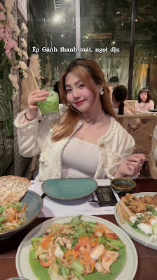 A woman is seated at a restaurant table in Thao Dien, Saigon, holding a green drink. The table features multiple dishes including a shrimp salad, a wr