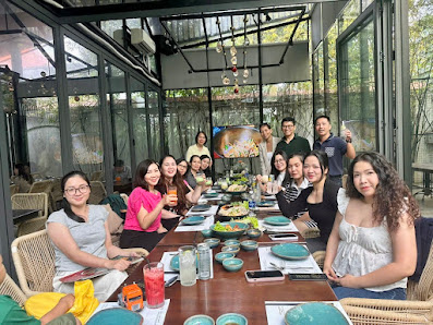 A group of people is gathered around a long dining table in an indoor setting that appears to be a restaurant or cafe, with large windows allowing nat
