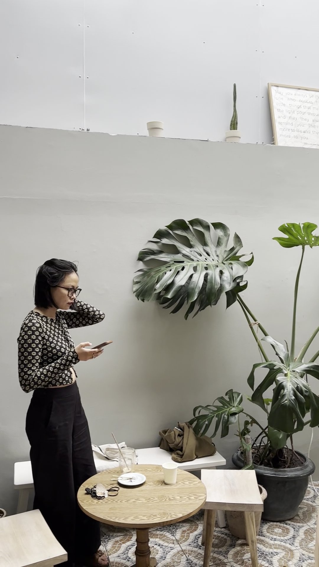 A woman in a black floral top and black pants stands near a round wooden table with a glass, cup, and small items. The background features a light gra