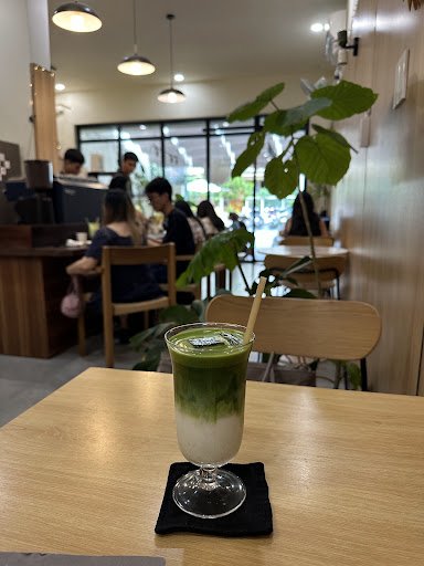 A green tea latte sits on a wooden table in an indoor cafe setting. The background shows patrons seated at tables, engaged in conversation or working 