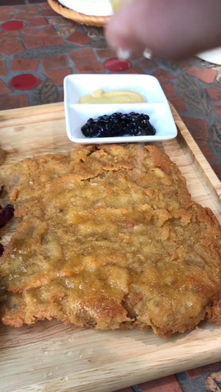 A close-up of a golden-brown fried dish on a wooden board, possibly a type of fritter or patty, with a small white bowl containing two condiments besi