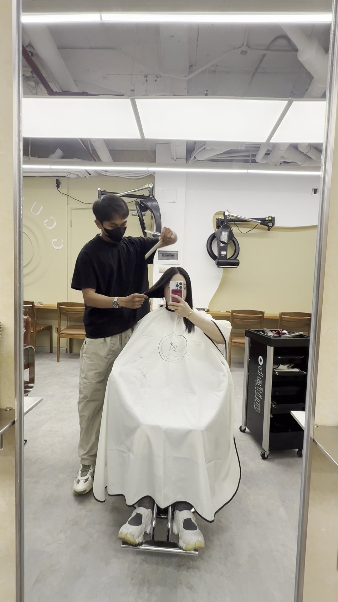 The image shows a hair salon interior where one person is getting their haircut by another individual who appears to be the stylist.