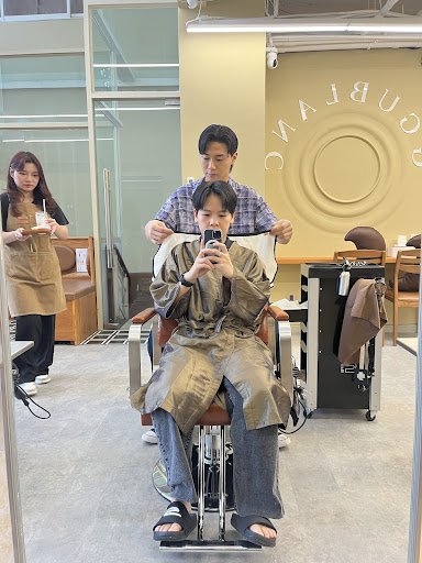 The image shows an interior view of a modern barbershop or hair salon named 'QUABRAVIE'. A customer is seated in the barber chair, wearing a brown cap