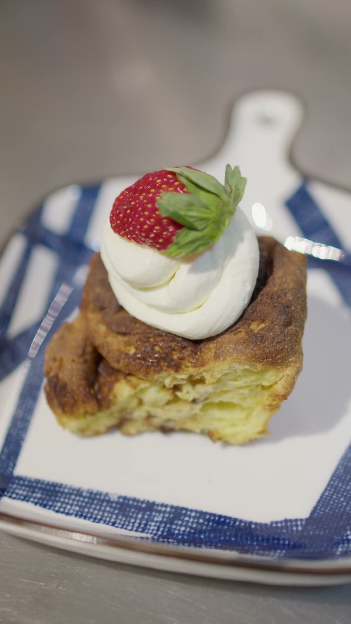 A plated dessert consisting of a golden-brown pastry topped with whipped cream and a strawberry garnish on a blue and white checkered plate.