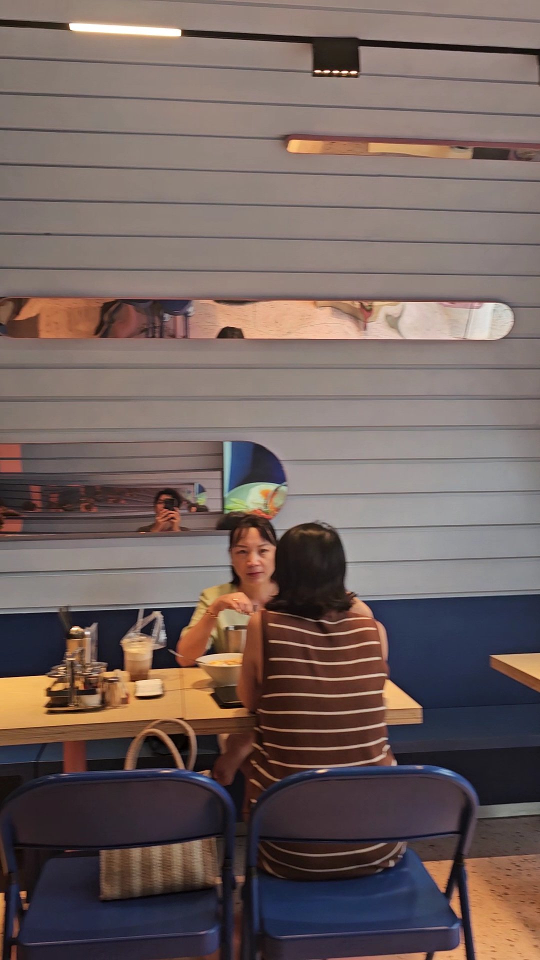 Two individuals are seated at a table in an indoor setting that appears to be a casual dining restaurant or cafe. The decor includes horizontal wooden