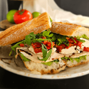 A sandwich cut in half and placed on a plate, featuring layers of bread, greens, red peppers, turkey or chicken slices, and possibly cheese.
