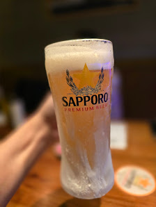 A close-up of a frothy beer glass labeled 'Sapporo Premium Beer'. The background is blurred but appears to be an indoor setting with warm lighting.