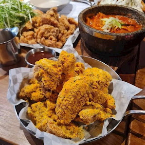 The image shows a close-up of golden-brown fried chicken pieces served in a metal bowl on paper towels with various dipping sauces beside it, along wi
