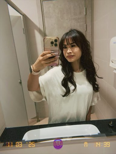 A woman is taking a selfie in front of a bathroom mirror while wearing a white top and holding a smartphone with a pink case.
