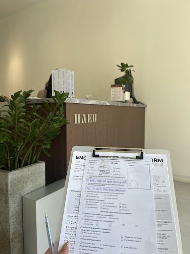 The image shows a reception area of an establishment named 'HAEI'. A clipboard with documents is being held in the foreground, and there's greenery on