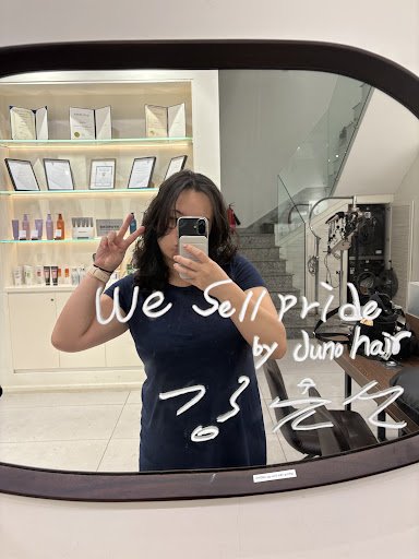 A woman is taking a selfie in front of a large mirror inside what appears to be a beauty salon or hairdresser's shop, with various products and framed