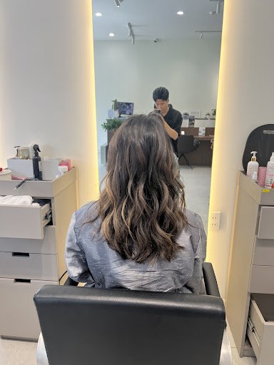 The image shows a salon interior where a person is seated in a chair receiving hair care services from another individual standing across the room, re