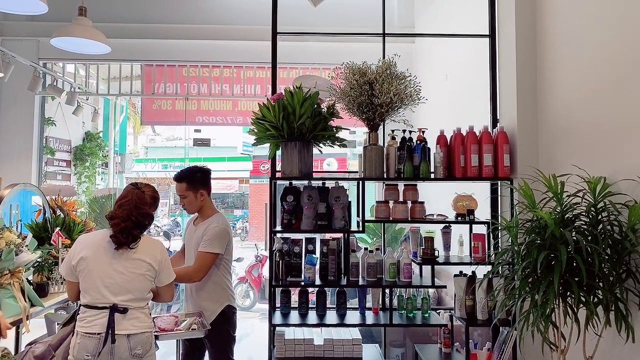 The image depicts an interior view of a beauty salon or hairdressing shop. A man and woman are interacting near the counter, which displays various pr
