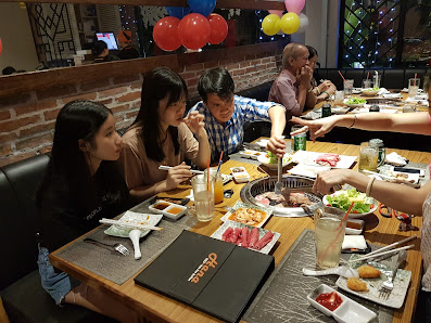 A group of people dining at a restaurant table with various dishes, drinks, and a central grill. The table features plates of food, a black menu, glas