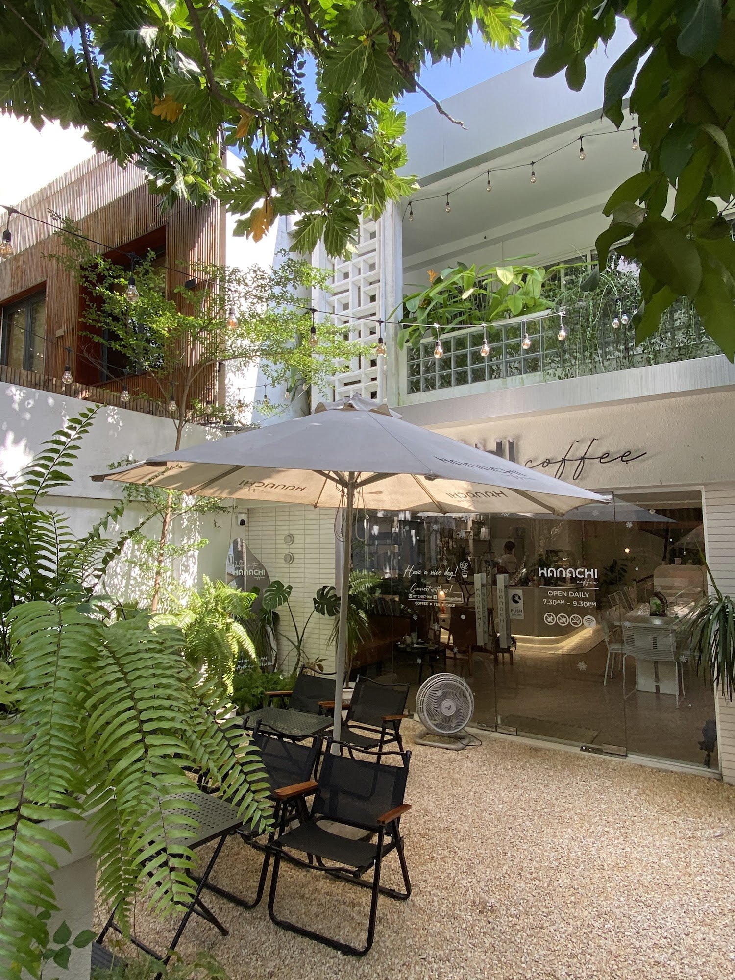 The image shows an outdoor seating area of a café named 'Hanaichi' located in Thao Dien, Saigon. The setting is modern and inviting with greenery surr