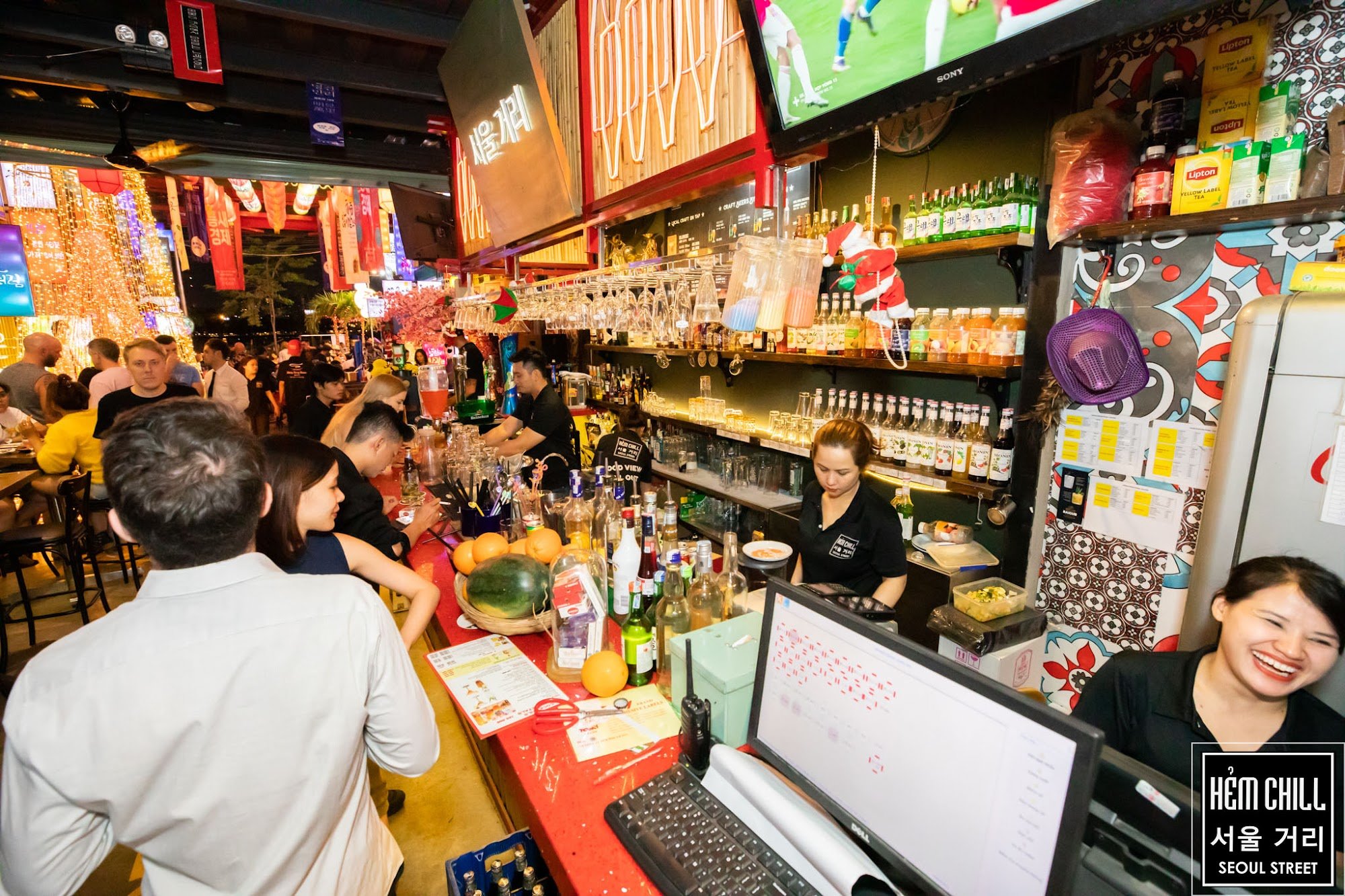 The image depicts a lively bar scene at night on Seoul Street in Thao Dien, Saigon. The interior is vibrant with colorful decorations and neon signs i