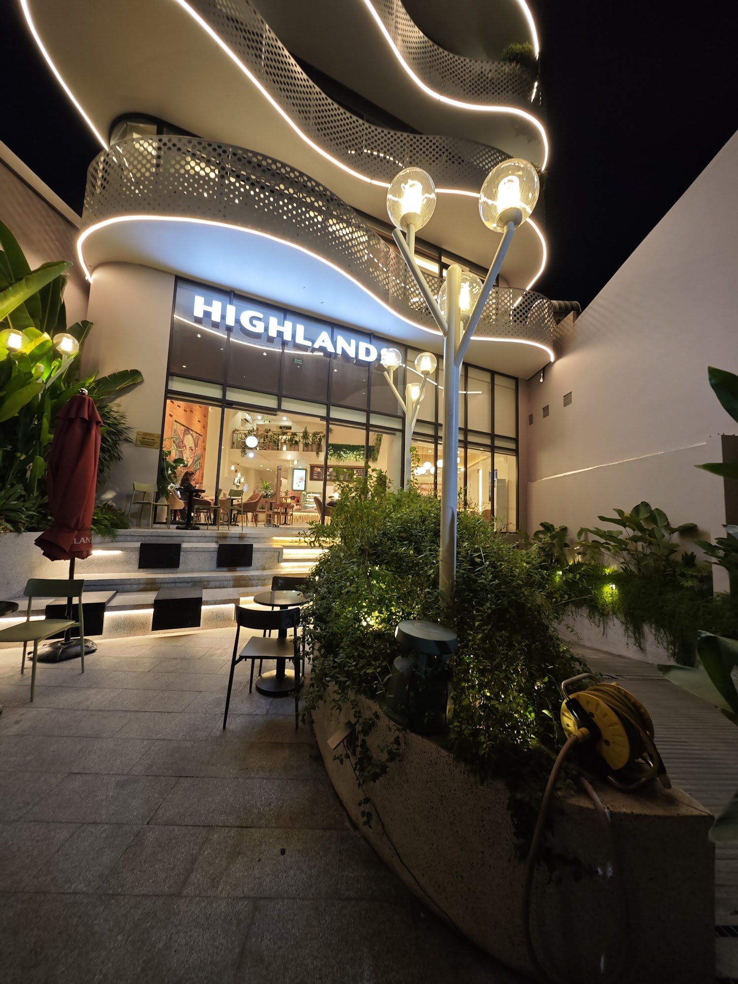 The image shows an exterior view of a modern café named 'Highland' at night time in Thao Dien, Saigon.