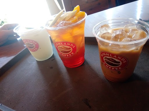 Three cups of beverages from Highlands Coffee on a tray, including what appears to be an iced coffee with ice cubes and milk in the foreground, a red-
