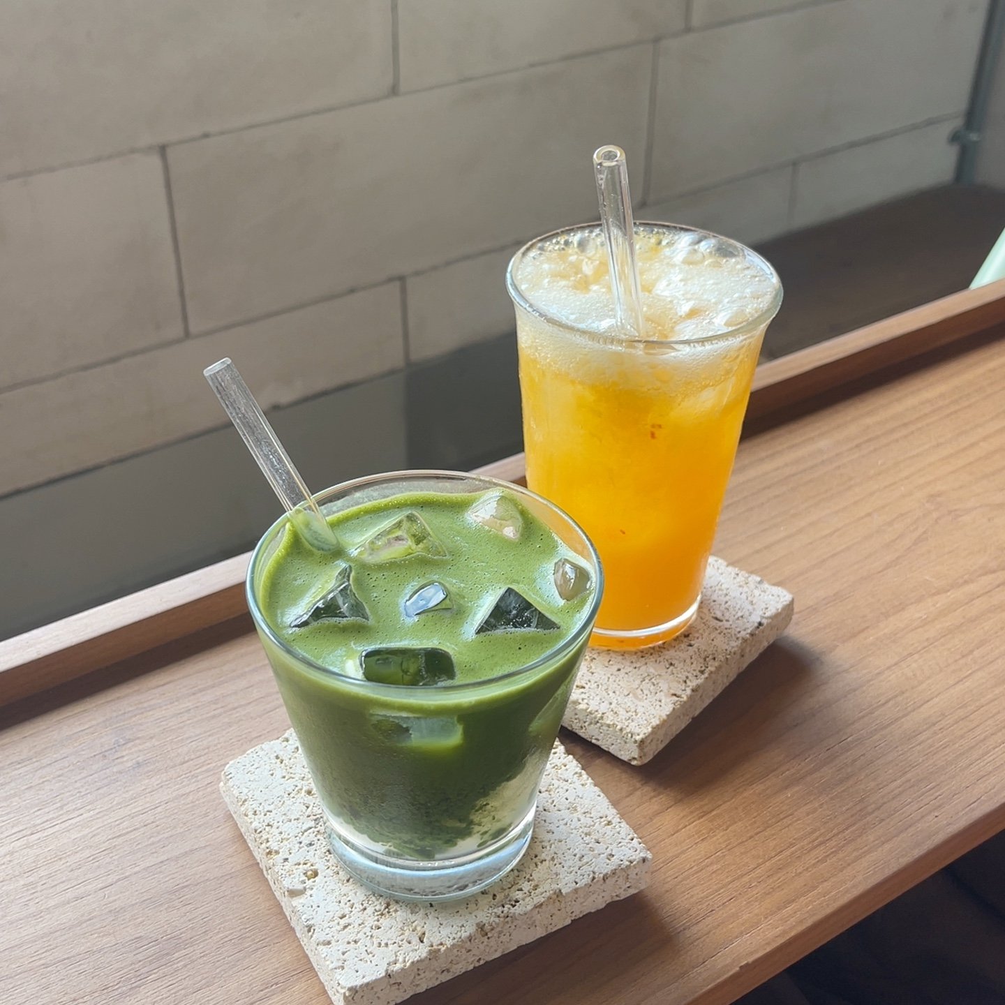 Two beverages are placed on a wooden surface; one is a green smoothie-like drink with ice cubes and the other appears to be an orange juice or similar