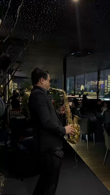 A man in a black suit playing a saxophone in a dimly lit indoor venue. The background features tables with chairs, other patrons seated, and large win