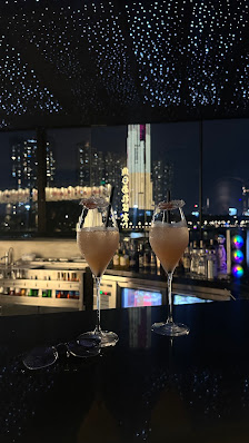 Two tall glasses filled with a light pink beverage, likely a cocktail or champagne, placed on a dark bar counter. The background features a nighttime 