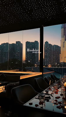 A modern restaurant interior with large windows overlooking a city skyline at sunset. Tables are set with glassware, plates, and rose petals, suggesti