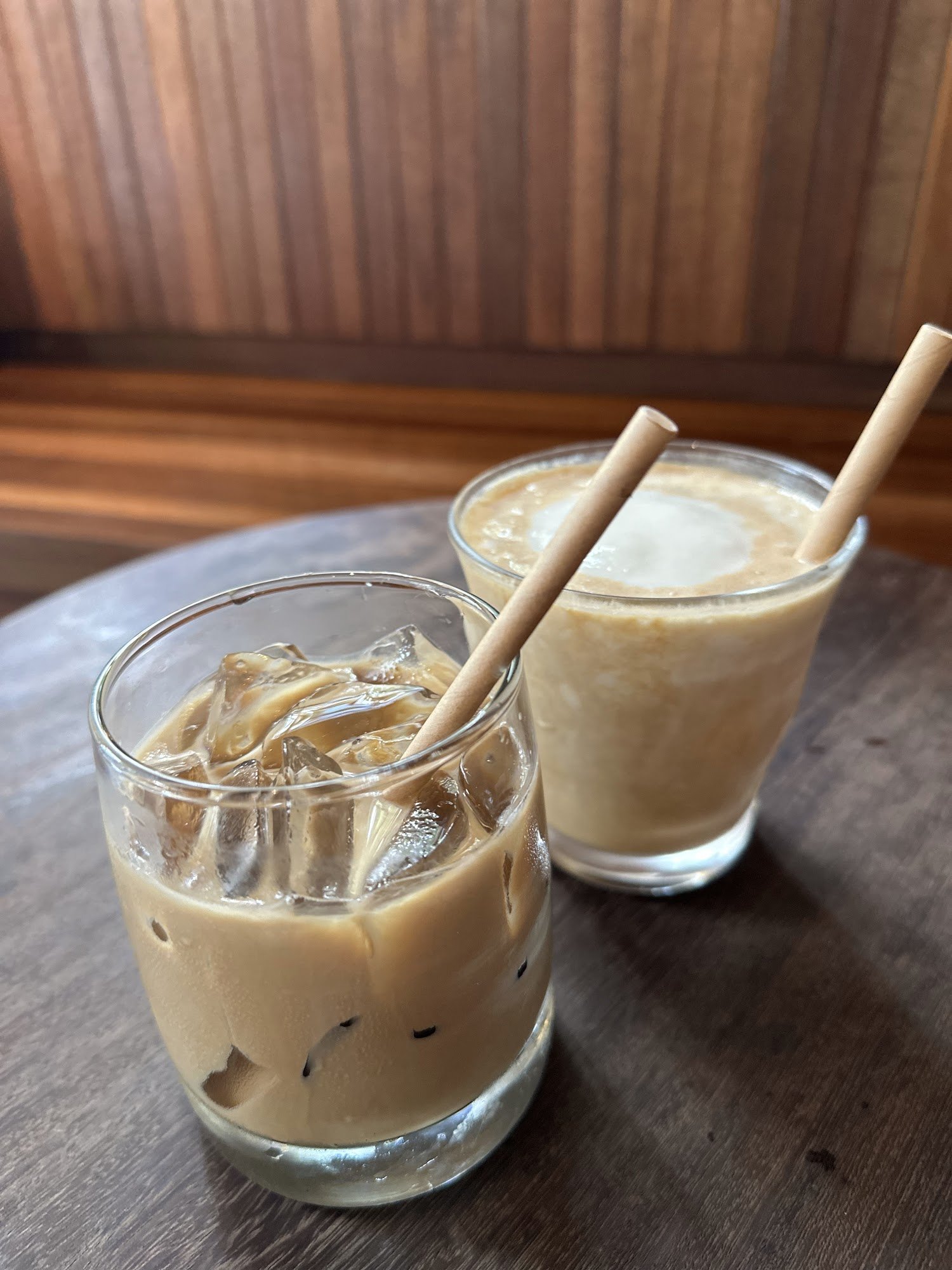 Two glasses of a creamy beverage topped with ice cubes and straws are placed on a wooden surface against a wood-paneled wall.