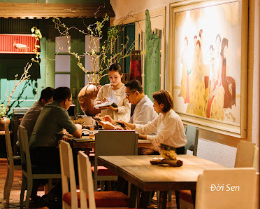A group of people dining at a restaurant table in Thao Dien, Saigon. The setting includes wooden tables, chairs with red cushions, a large framed artw