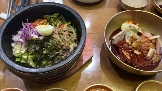 The bowls contain Korean side dishes known as banchan and a spicy noodle soup called naengmyeon. The ingredients visible include kimchi, seaweed, sliced radish, boiled egg, meat, vegetables, noodles, and various sauces.