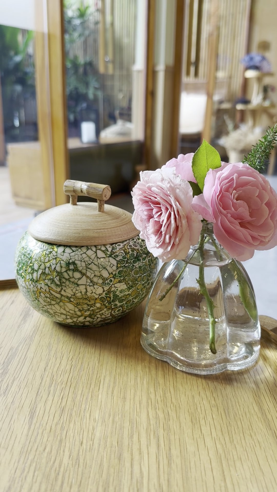 A wooden table features a decorative ceramic jar with green and gold crackle glaze and a bamboo lid, alongside a clear glass vase holding pink peonies