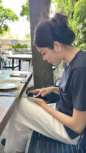 A woman with dark hair tied up, wearing a dark gray t-shirt and white pants, is seated at an outdoor table in a restaurant. The table has plates, cutl