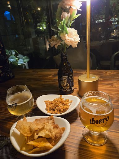 The image shows a cozy dining setting at Thao Dien, Saigon featuring a table with various dishes and drinks including beer in branded glasses, bowls o