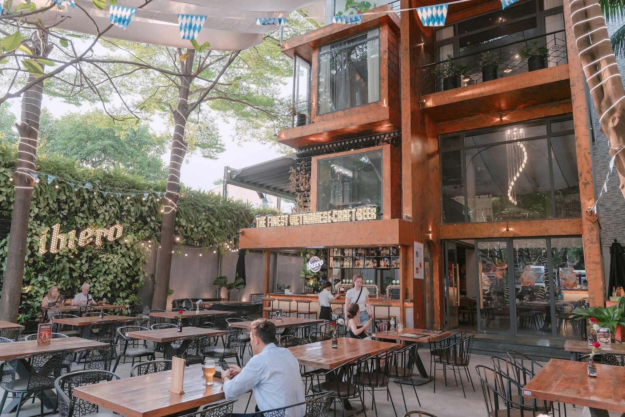 The image depicts an outdoor seating area of a venue in Thao Dien, Saigon. It features wooden tables and black metal chairs with people dining and soc