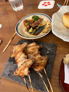 The image shows a plate of skewered grilled meat on a dark slate board accompanied by what appears to be pickled vegetables and possibly some sauce or