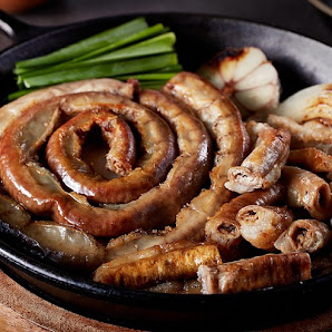 A close-up of a dish featuring various types of grilled sausages and offal in a black pan with green onions on the side.