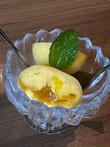 A dessert dish served in a clear glass bowl on a wooden table. The dessert appears to be a yellow custard with a green mint leaf garnish, and it is ac