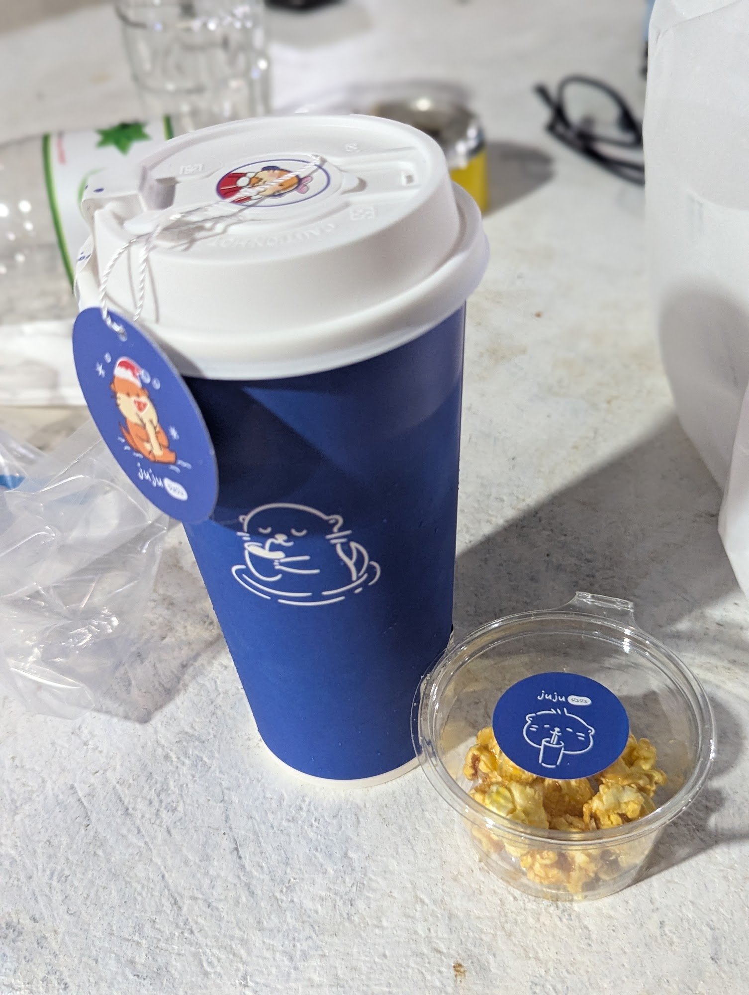 A blue reusable coffee cup with a white lid and a cartoon character design on the side is placed next to a clear plastic container filled with popcorn