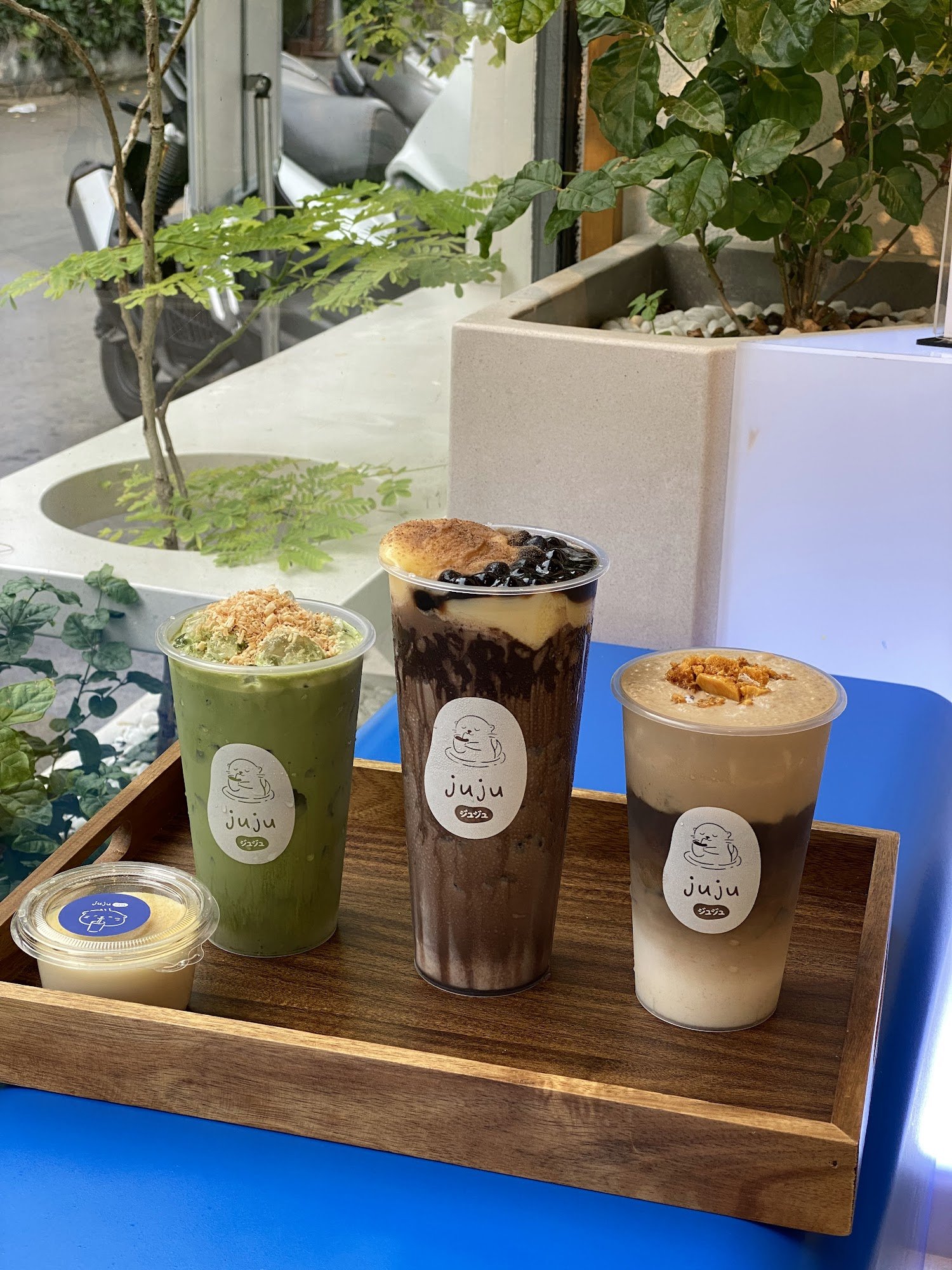 A wooden tray holds four beverages from a café named 'juju', featuring various flavors and toppings such as shredded coconut and black sesame seeds.