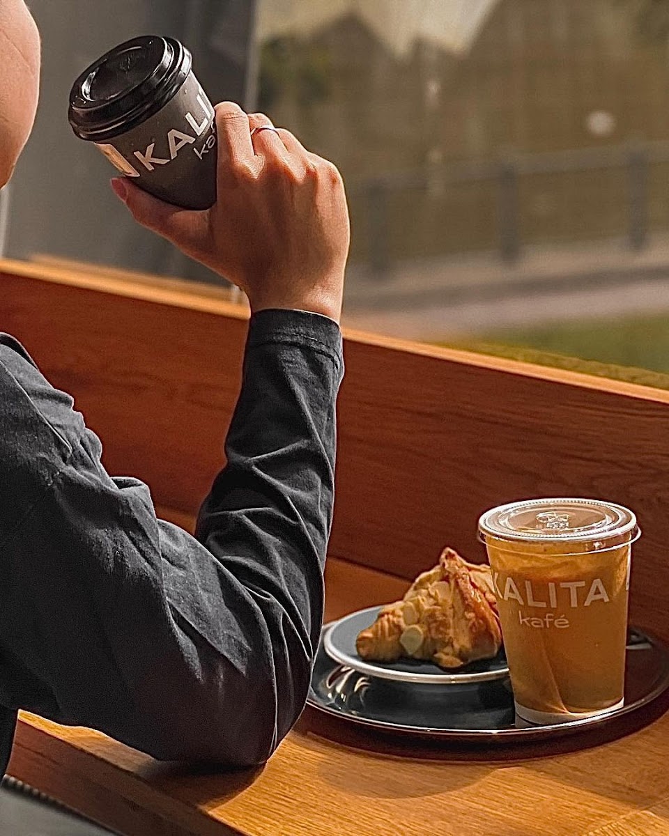 A person is seated at a wooden table in an indoor setting, enjoying a coffee from 'KALITA' with their right hand while holding the cup close to their
